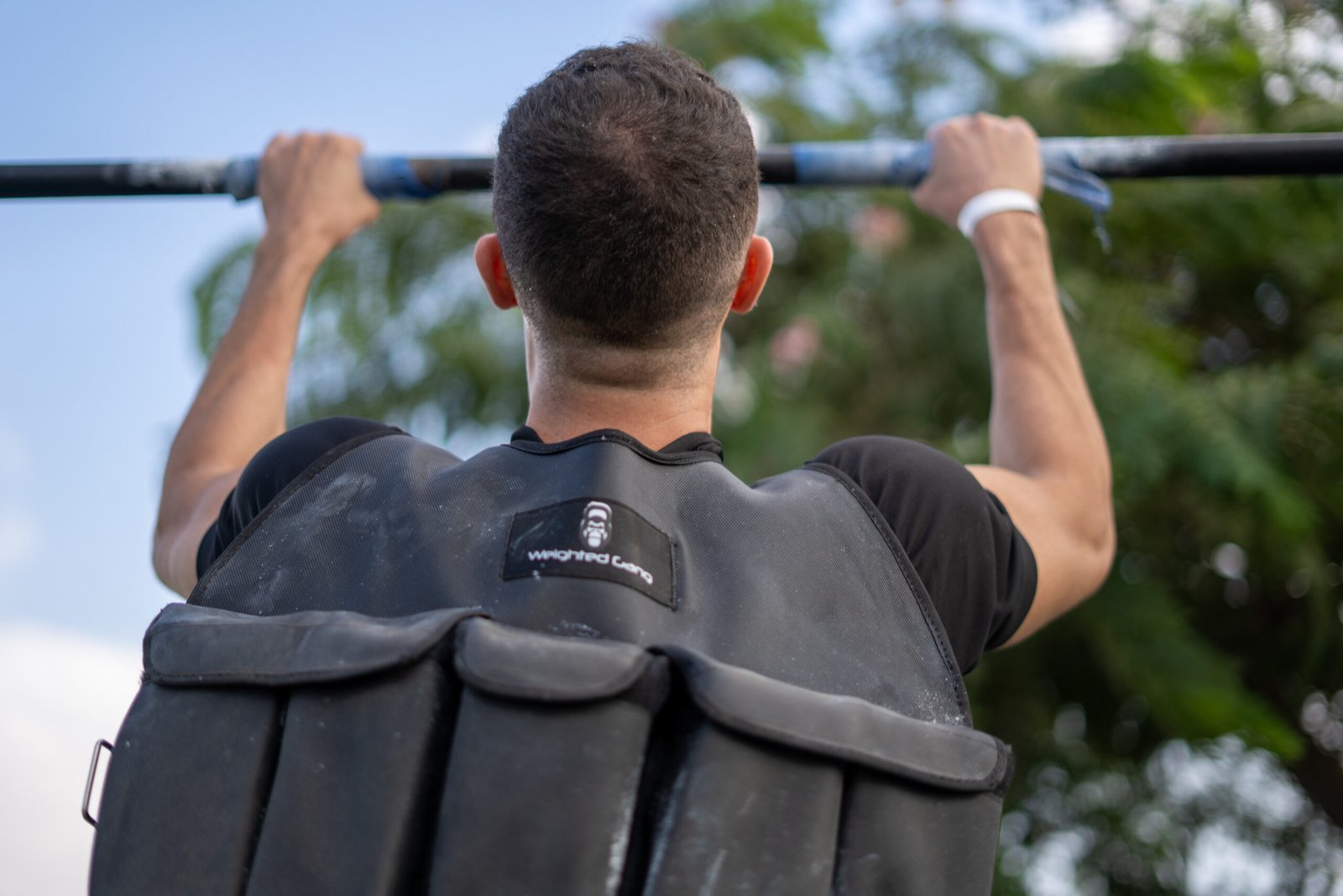 Man with weighted vest doing pull-up outdoors with trees in background.